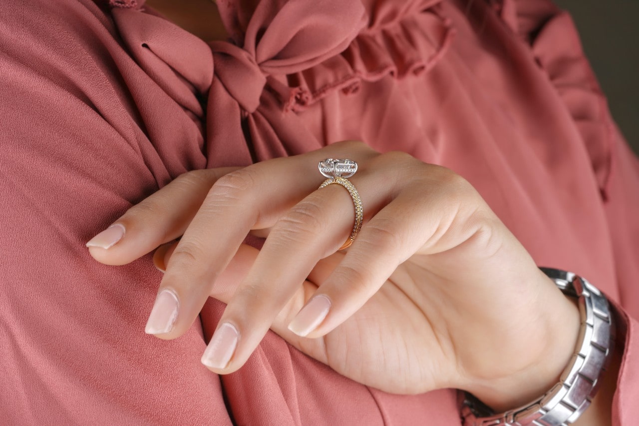 A hand with manicured nails rests against a pink blouse, showcasing a sparkling ring.