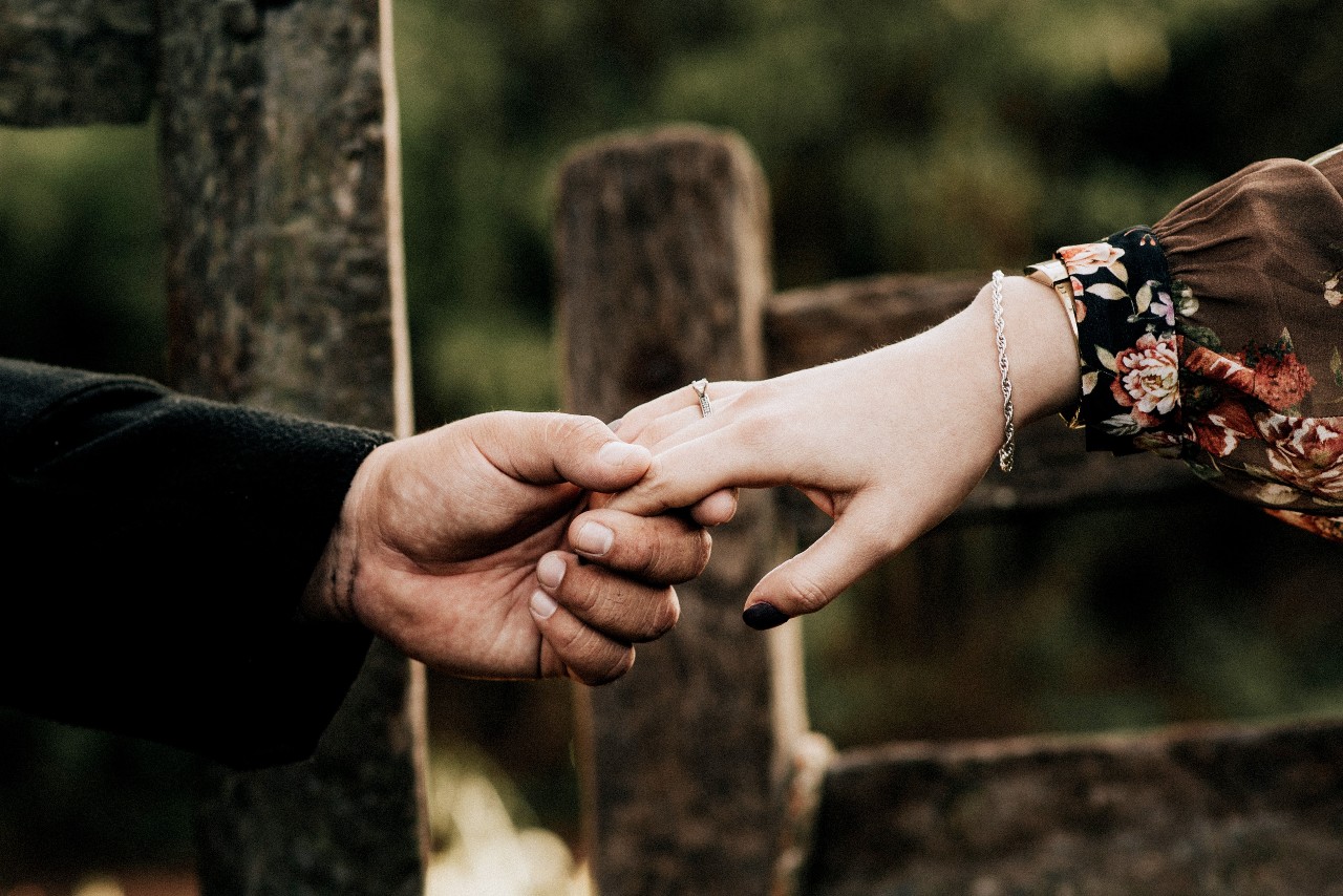 Close-up of a couple holding hands gently in front of a rustic wooden fence with a strong emphasis on the woman's beautiful ring.