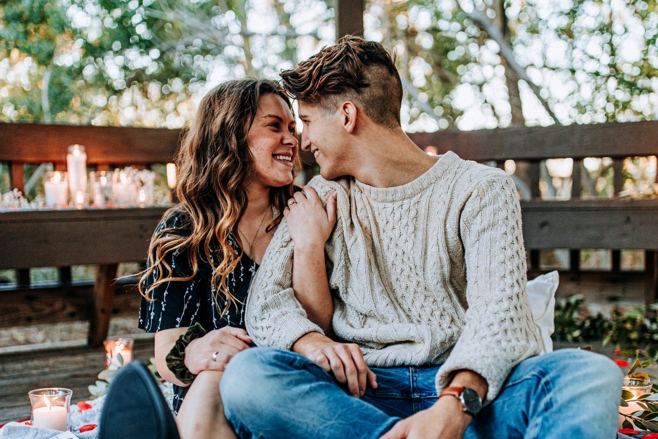 a couple sitting on the floor of a wooden structure outdoors, smiling at one another