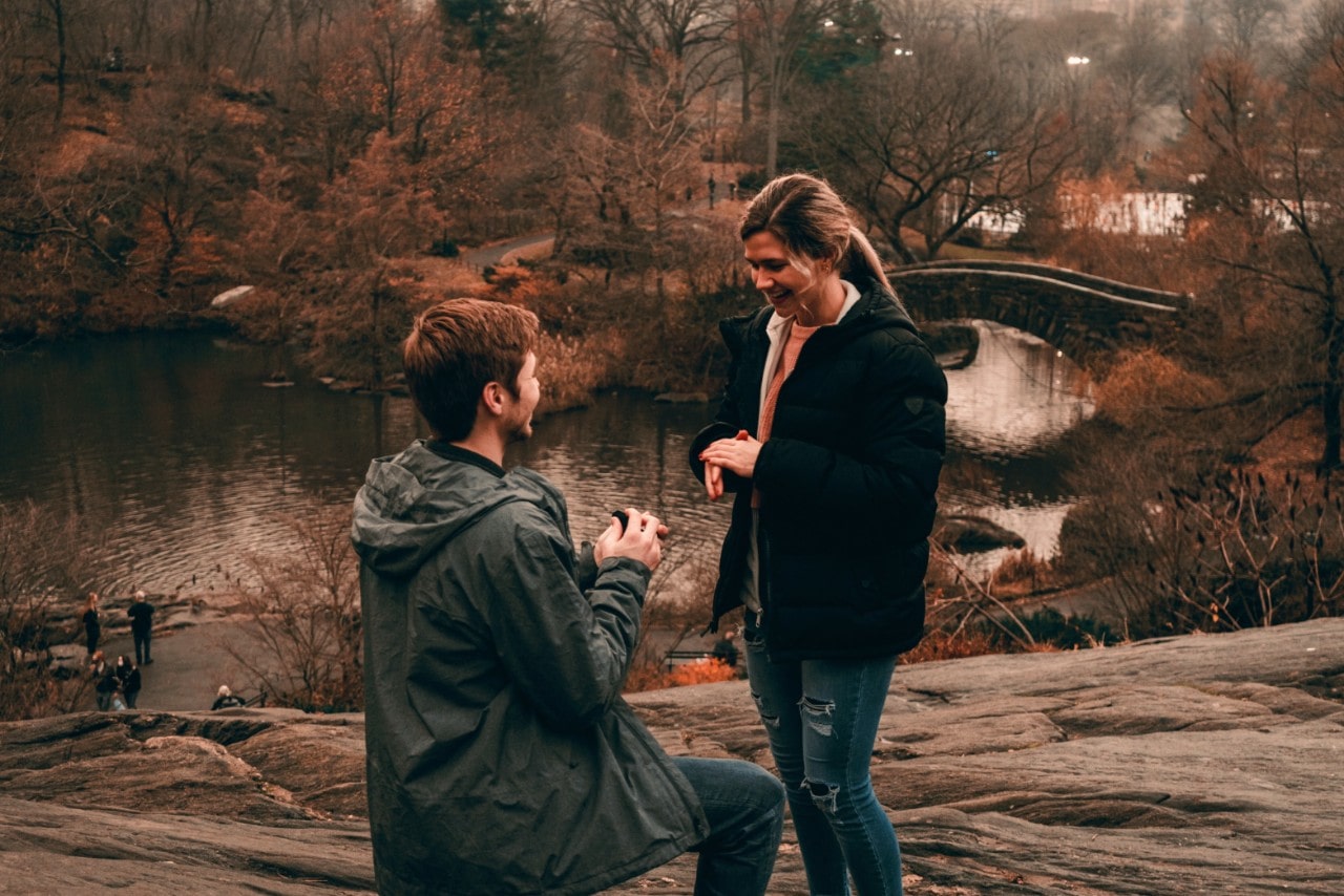 a man on one knee holding a ring box in front of a woman next to a river in the fall