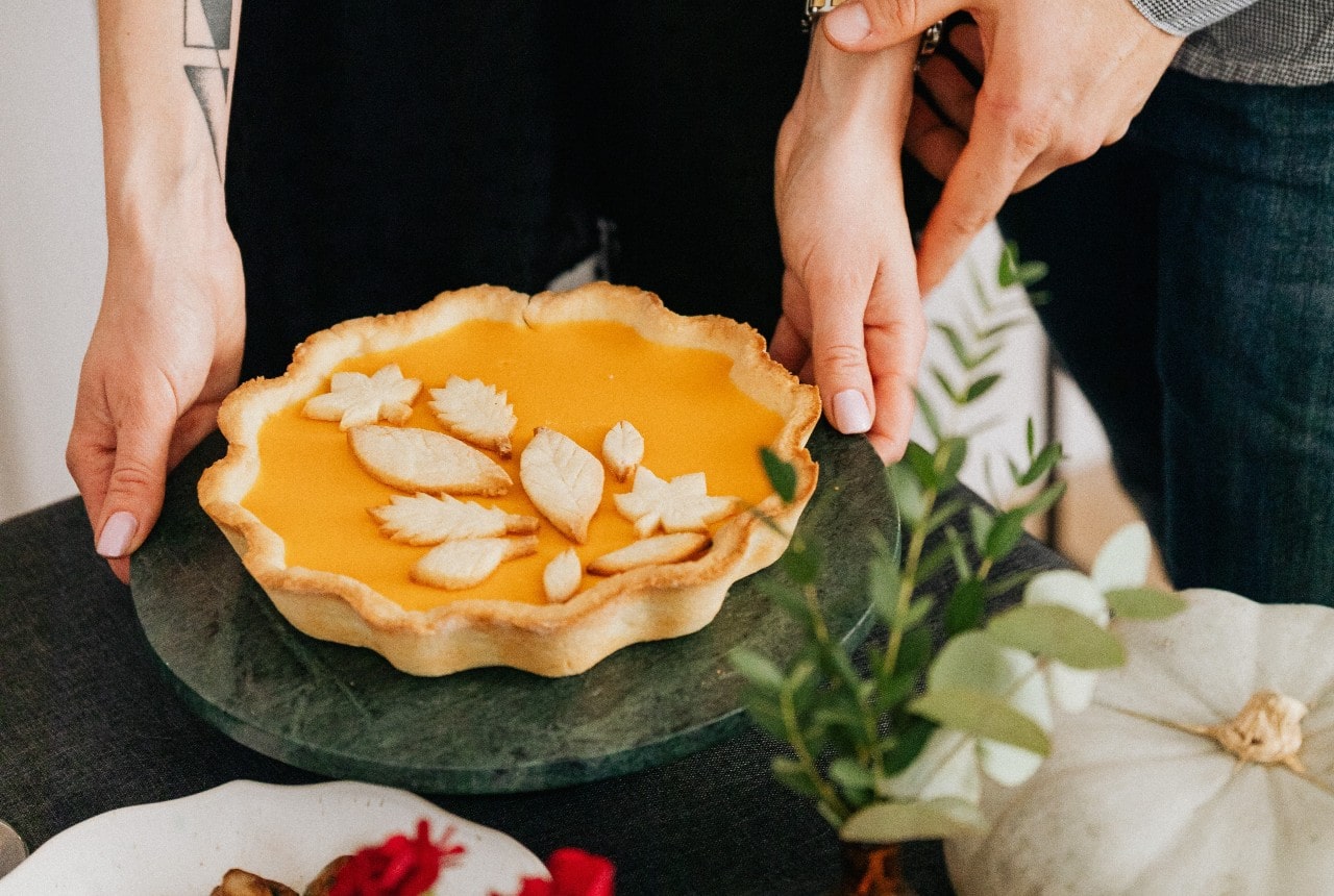 A couple puts a decorated pumpkin pie on the edge of a fall-adorned table.