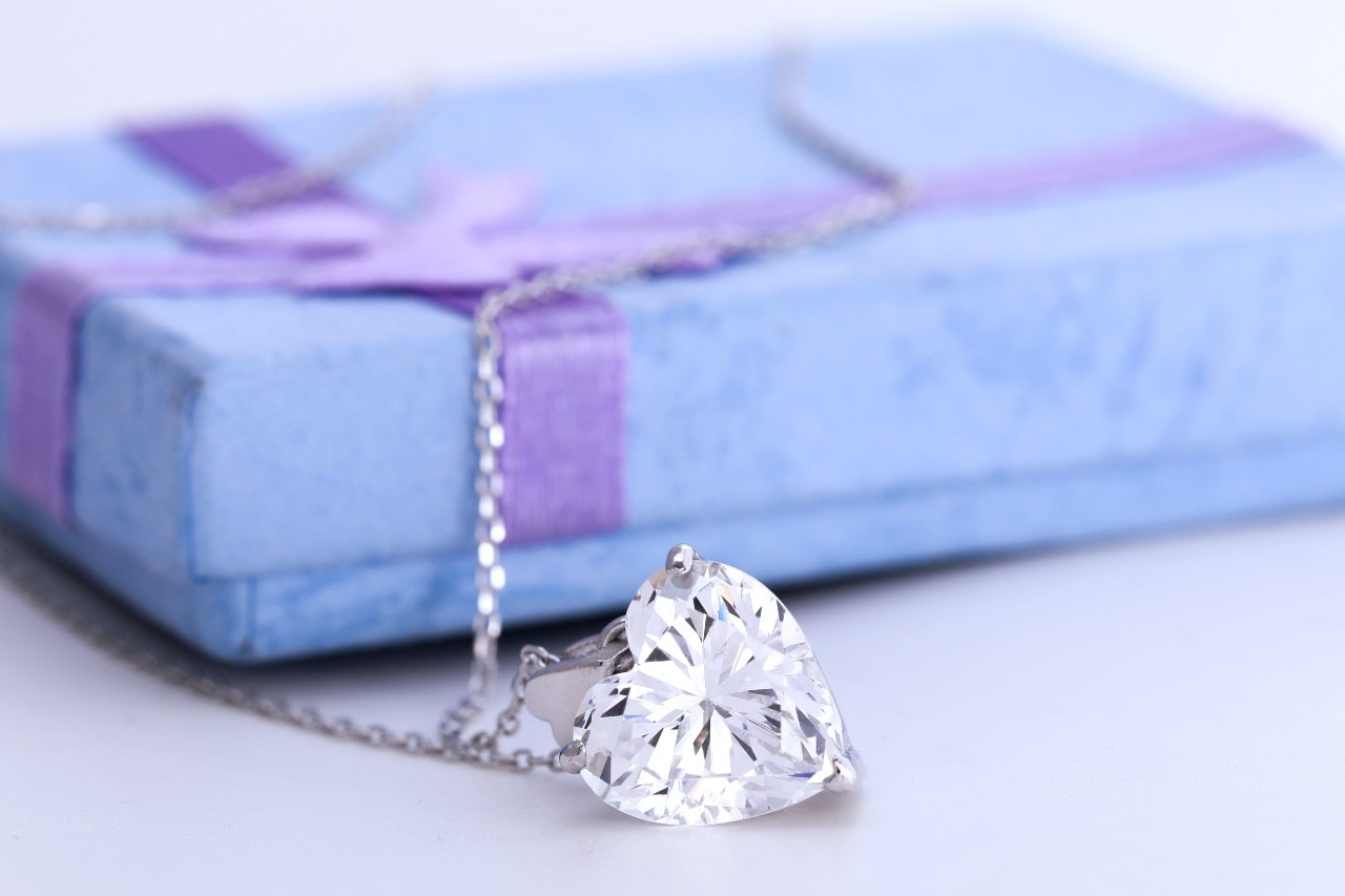 A heart-shaped diamond necklace laid atop a blue gift box with a purple ribbon.