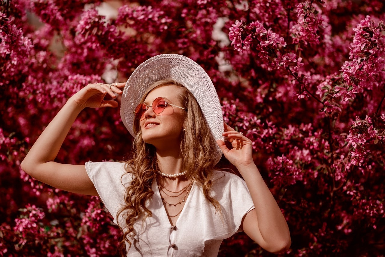 A woman adjusts her sunhat in front of a tall, blooming bush.