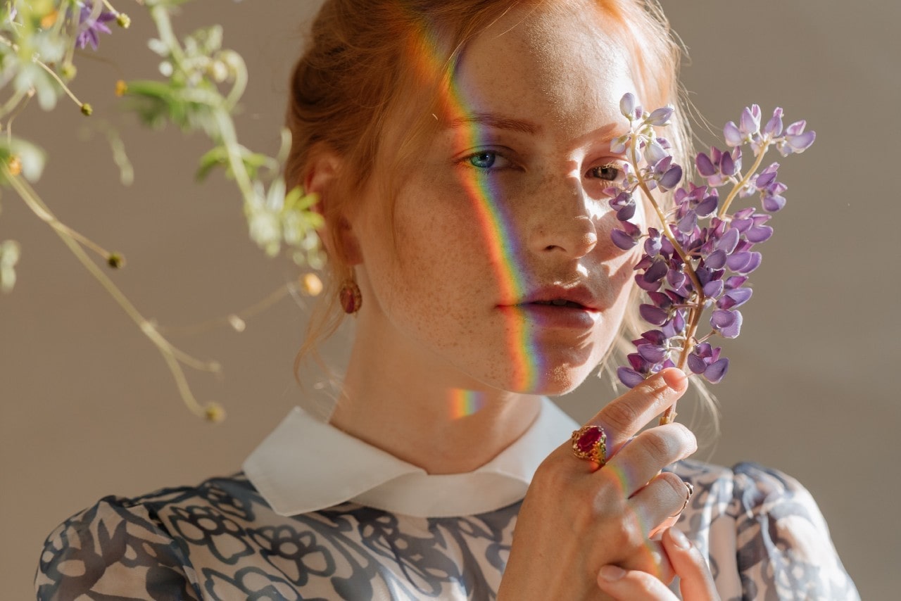 A styled woman with a rainbow across her face holds a purple flower to her face.