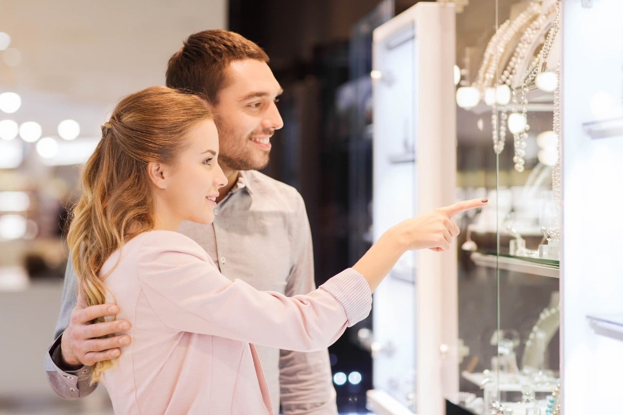 Smiling couple browsing jewelry; woman points at a display while her partner stands behind her, hand gently resting on her back.