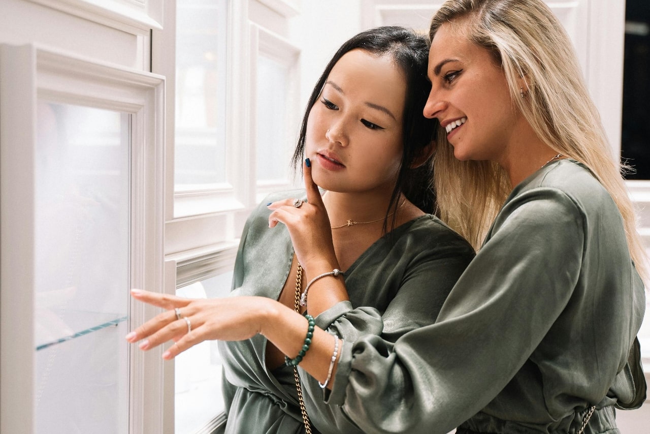 Two women in green outfits smile and point at a jewelry display in a luxurious boutique.