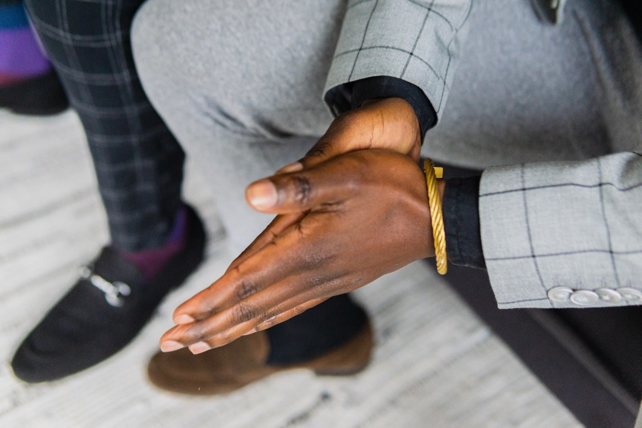A top-down view of a well-dressed man in a suit sitting in a chair, a gold bracelet on his left wrist.