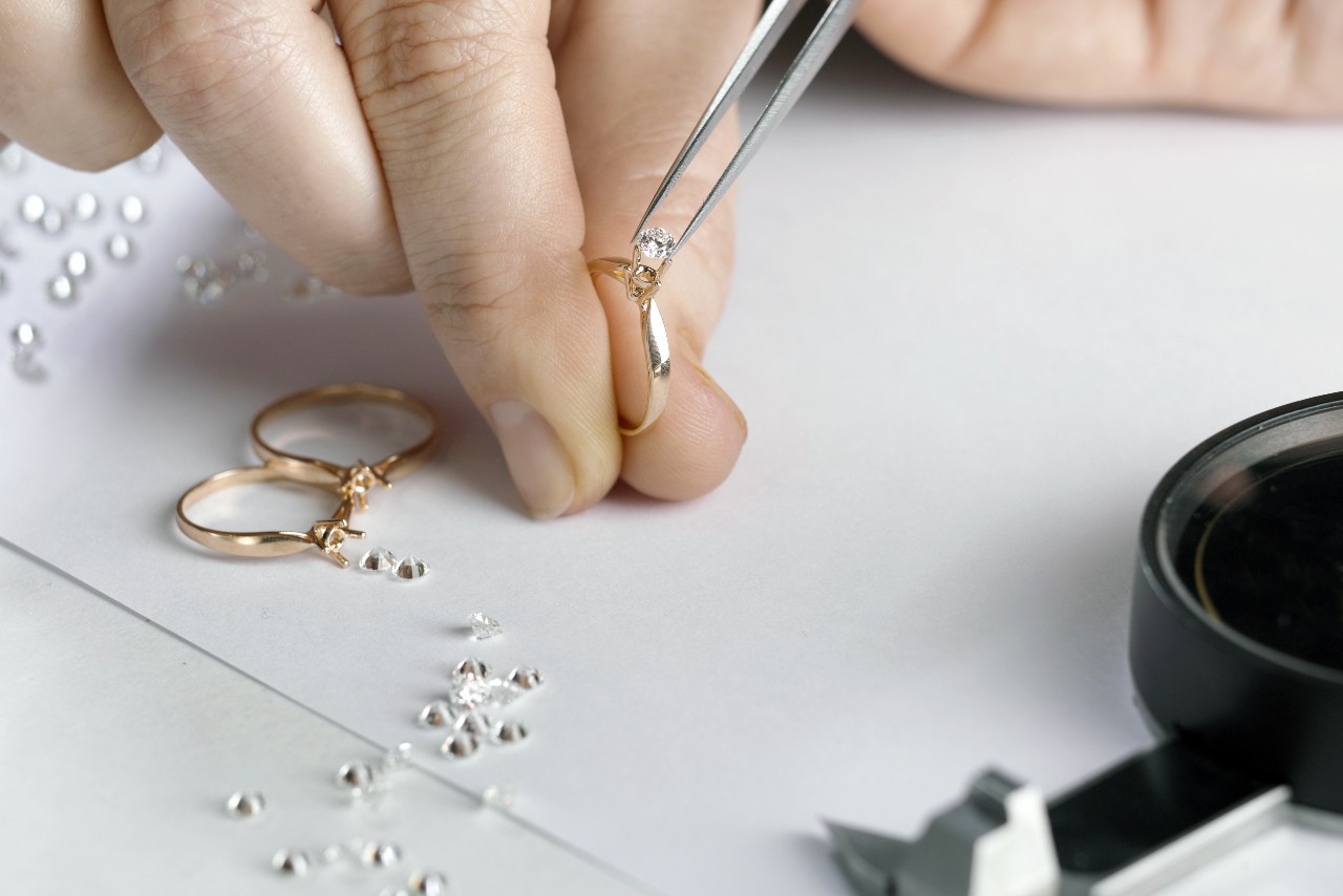 A close-up of a jeweler’s hand setting a diamond into a gold ring with tweezers, surrounded by loose diamonds and two additional ring settings on white paper.