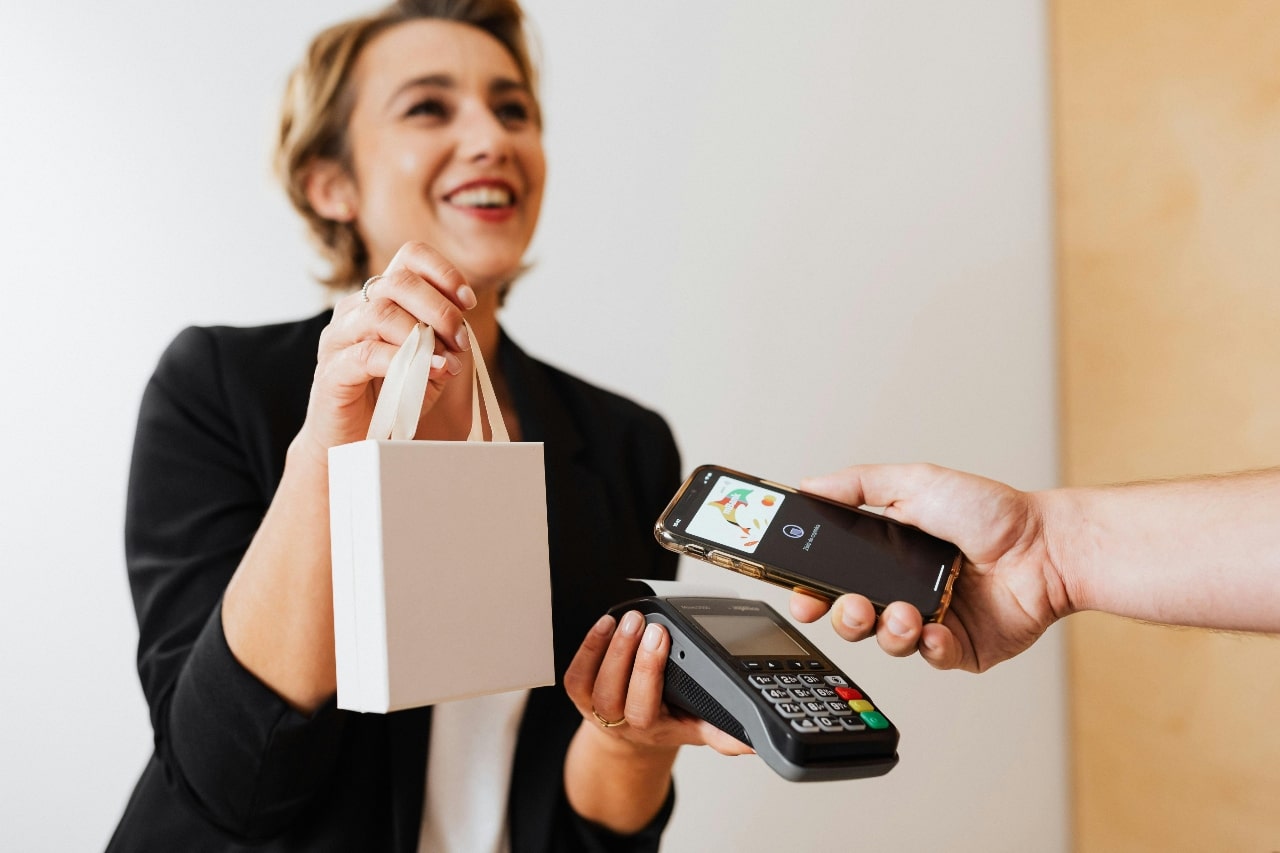 A smiling woman in a black blazer hands a small white shopping bag to a customer paying with a smartphone over a card reader, conveying a cheerful, modern shopping experience.