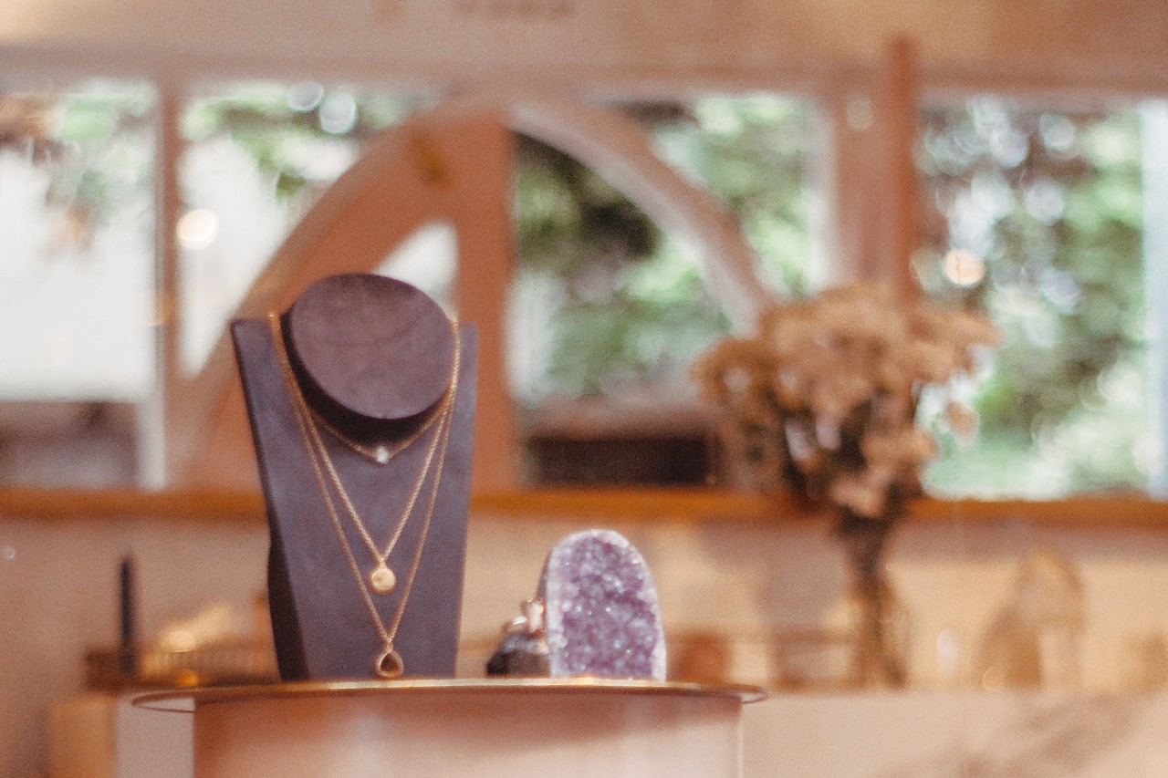 A layered gold necklace is displayed on a black jewelry stand beside a purple crystal inside a boutique with blurred greenery in the background.