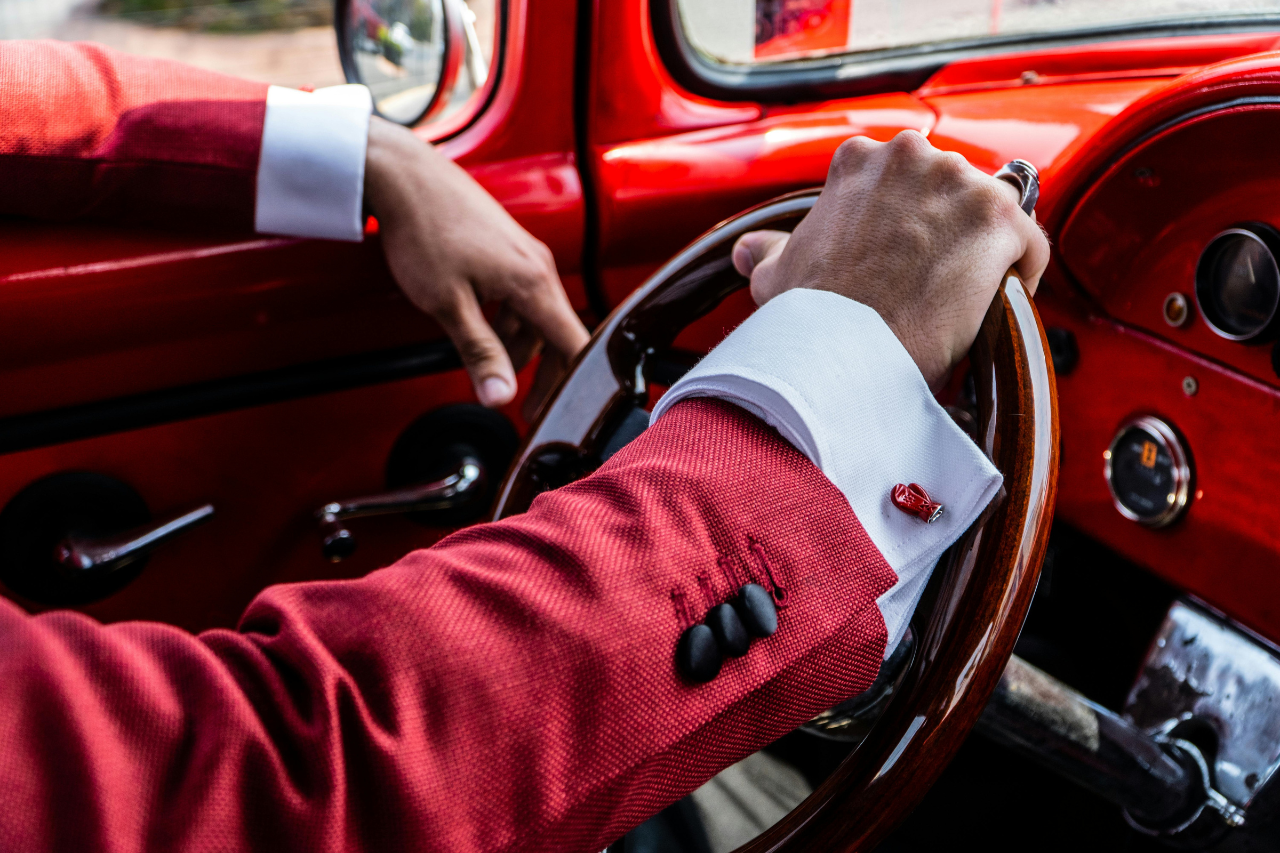 A close-up of a man’s arms as he drives a luxury car, wearing a red suit with boxing glove-shaped cufflinks. A close-up of a man’s arms as he drives a luxury car, wearing a red suit with boxing glove-shaped cufflinks.