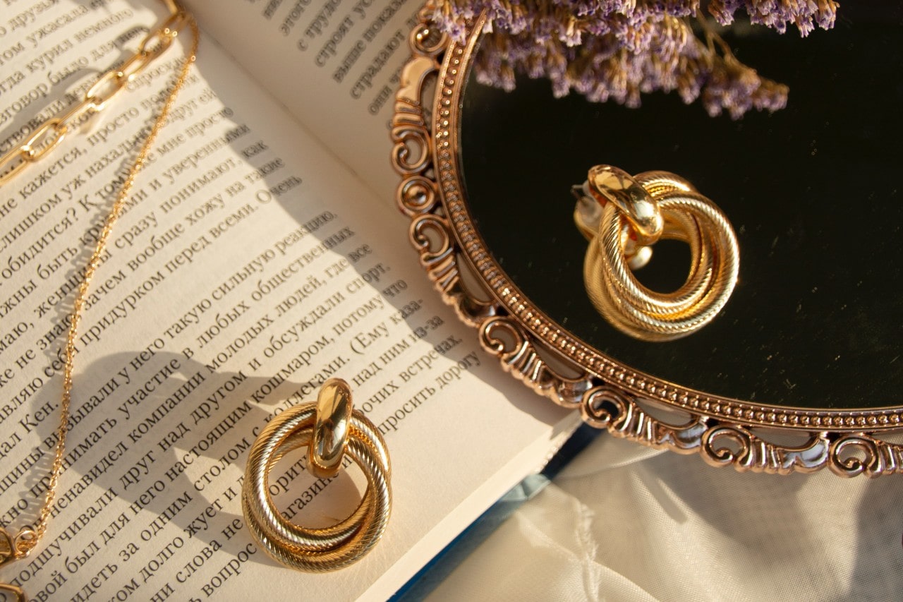 a pair of gold earrings resting on an open book and a mirror.