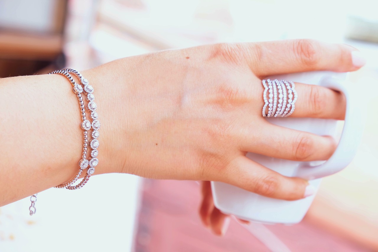 A hand adorned with a sparkling diamond bracelet and a multi-row diamond ring gracefully holds a white cup.