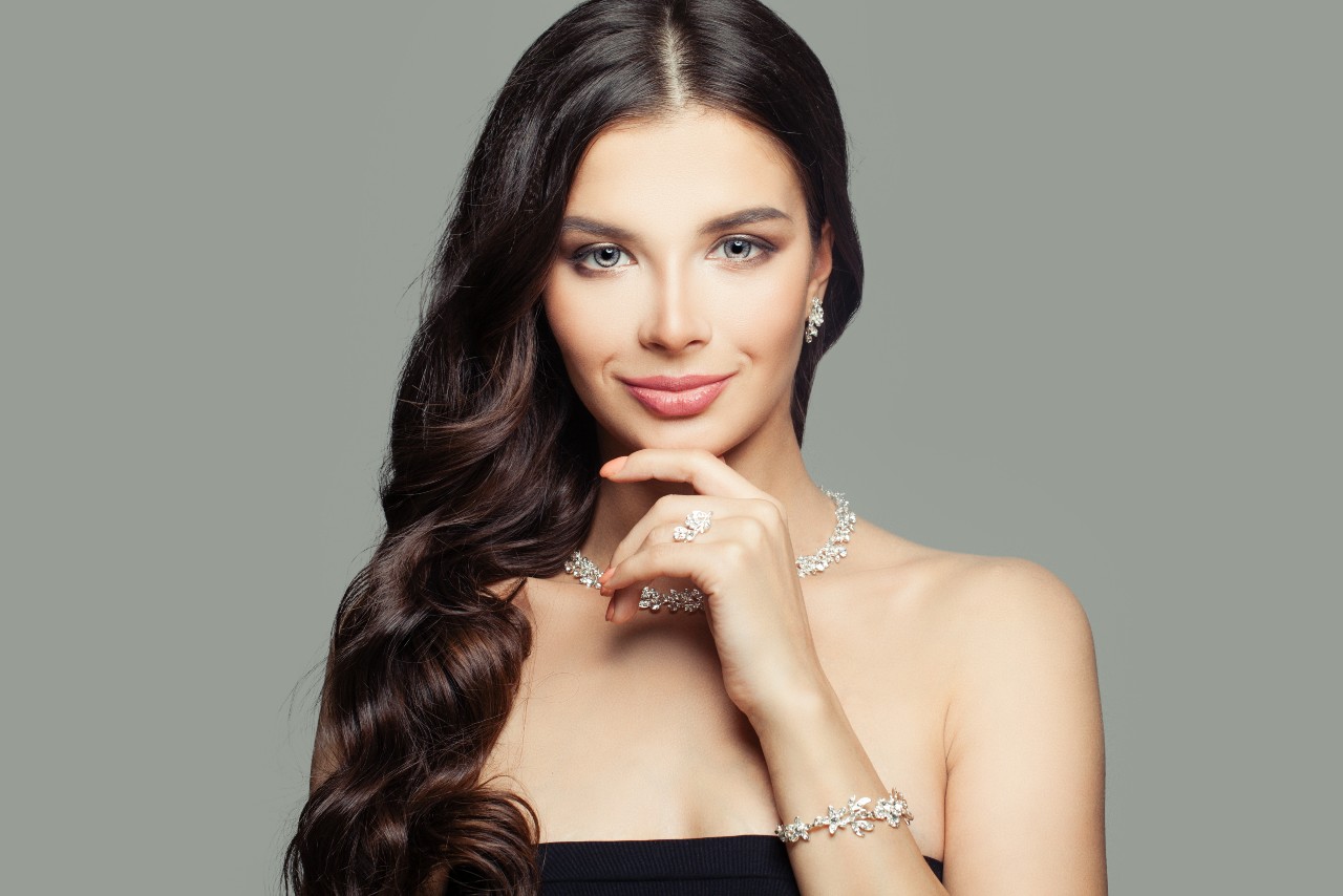 A woman with long dark hair poses elegantly wearing a matching diamond necklace, earrings, bracelet, and ring set.