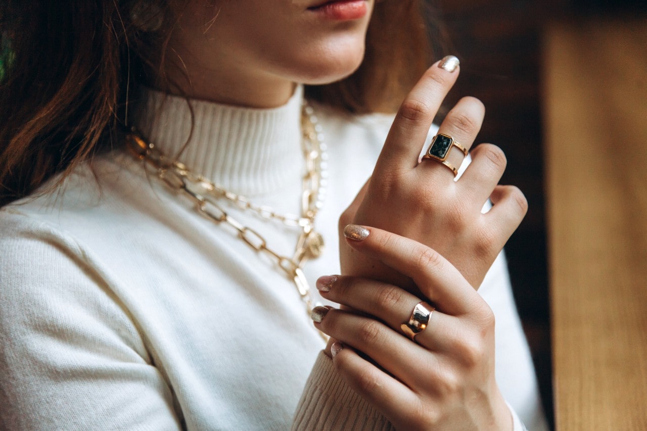 A close up of a woman wearing a white turtleneck and chunky yellow gold jewelry, layered chain necklaces and fashion rings
