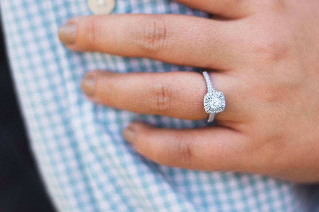 A close up of a hand resting atop a blue gingham shirt, a white gold diamond halo engagement ring on the ring finger.
