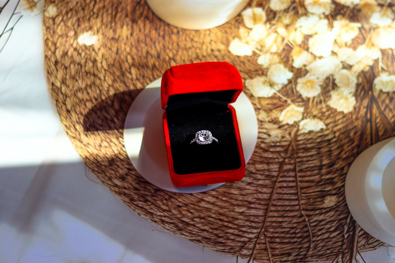 A white gold halo diamond engagement ring inside a red jewelry velvet box, displayed on top of a woven mat.