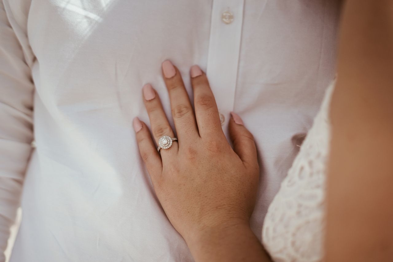 A close-up of a bride’s hand, adorned with white gold double halo diamond engagement ring, resting on her groom’s chest.