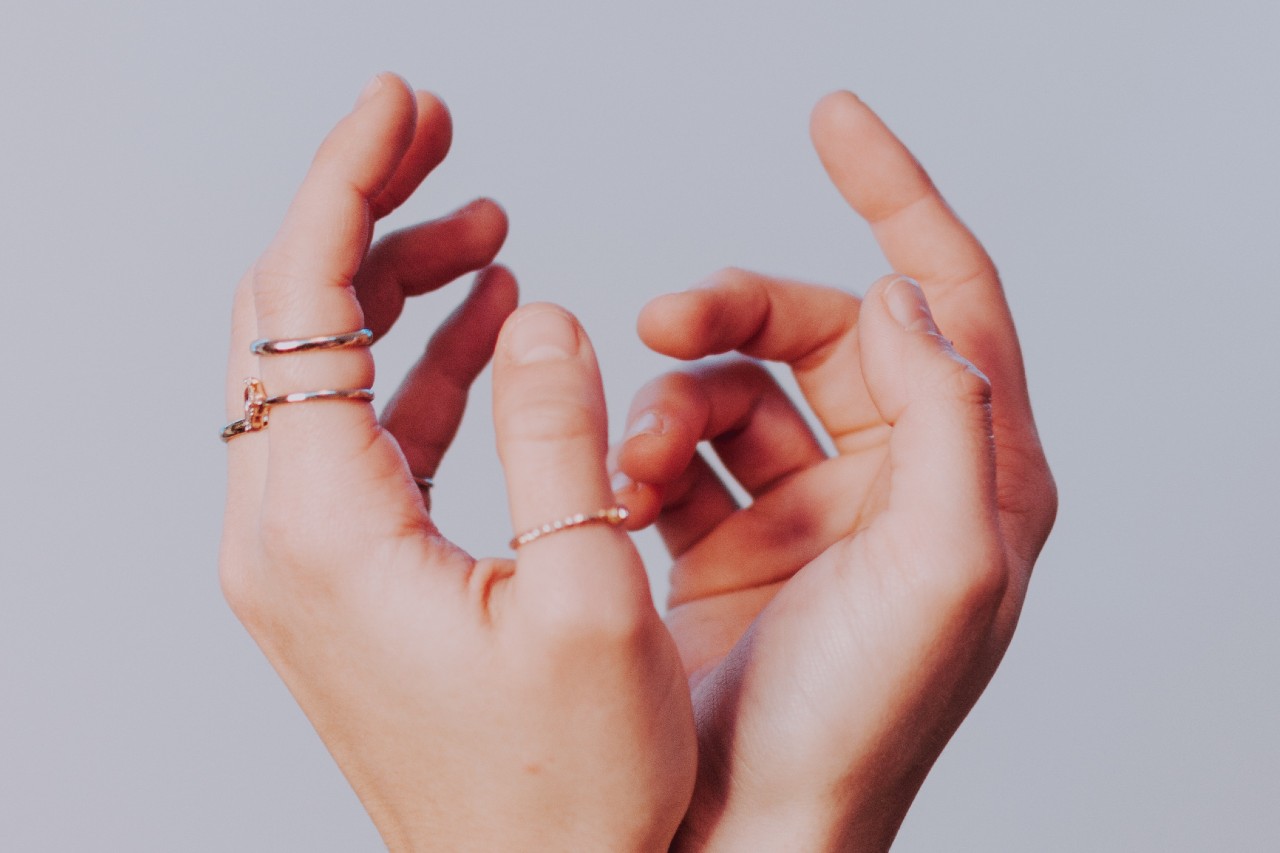 A pair of hands wearing thin gold rings with minimalist designs, posed against a soft pastel background