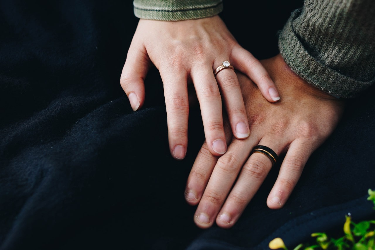 A woman resting her hand on top of her husband's hand, showing off their wedding rings against a dark navy fabric background.