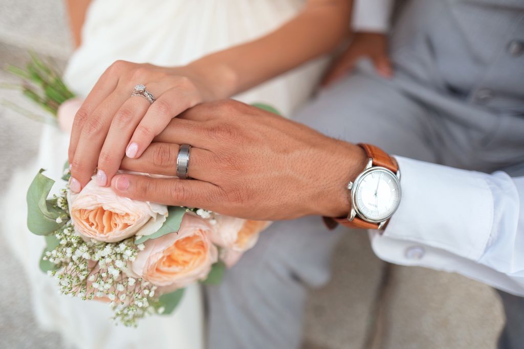 A freshly married couple placing their hands on top of one another’s and their bouquet of pink peonies, showcasing their wedding rings and a silver watch.