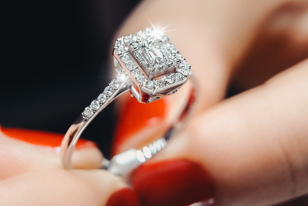 a woman with red nails holds an emerald-cut diamond halo ring under a light.