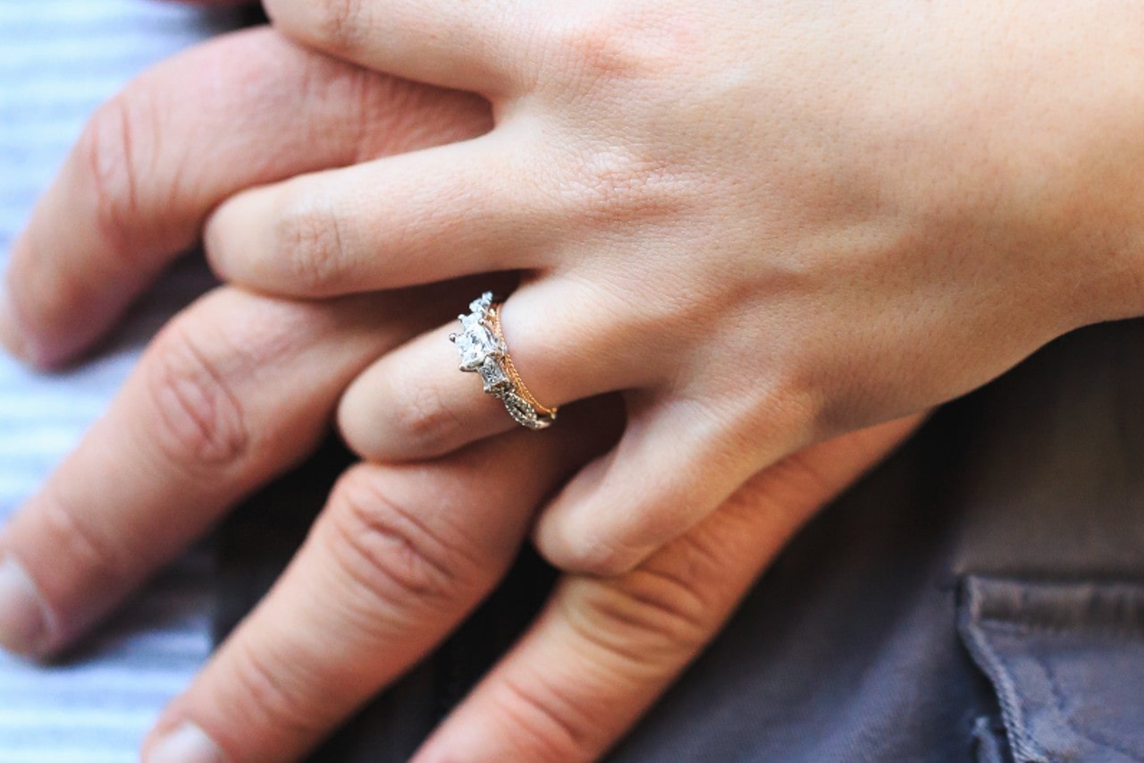 A couple holding hands, showing a three-stone engagement ring. A couple holding hands, showing a three-stone engagement ring.