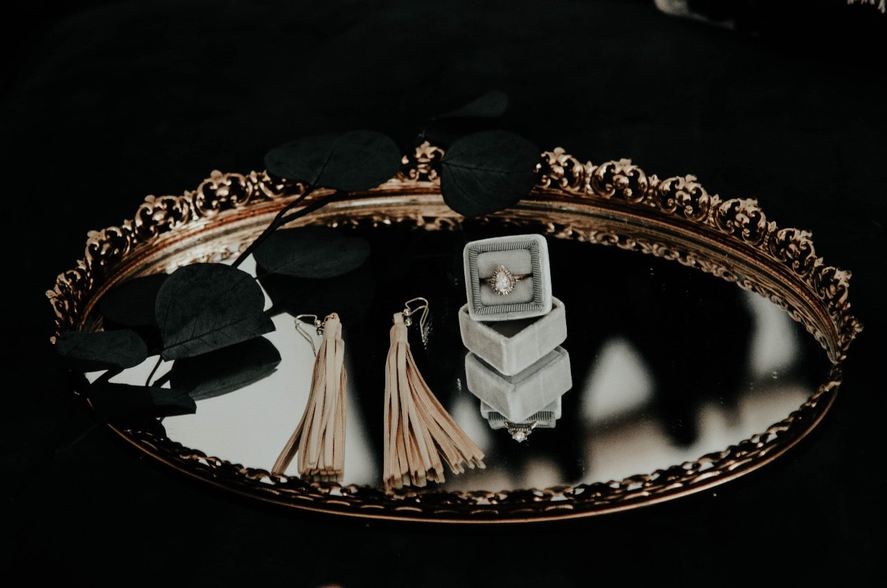 An engagement ring in a box sits on a tray with tassel earrings and black leaves.