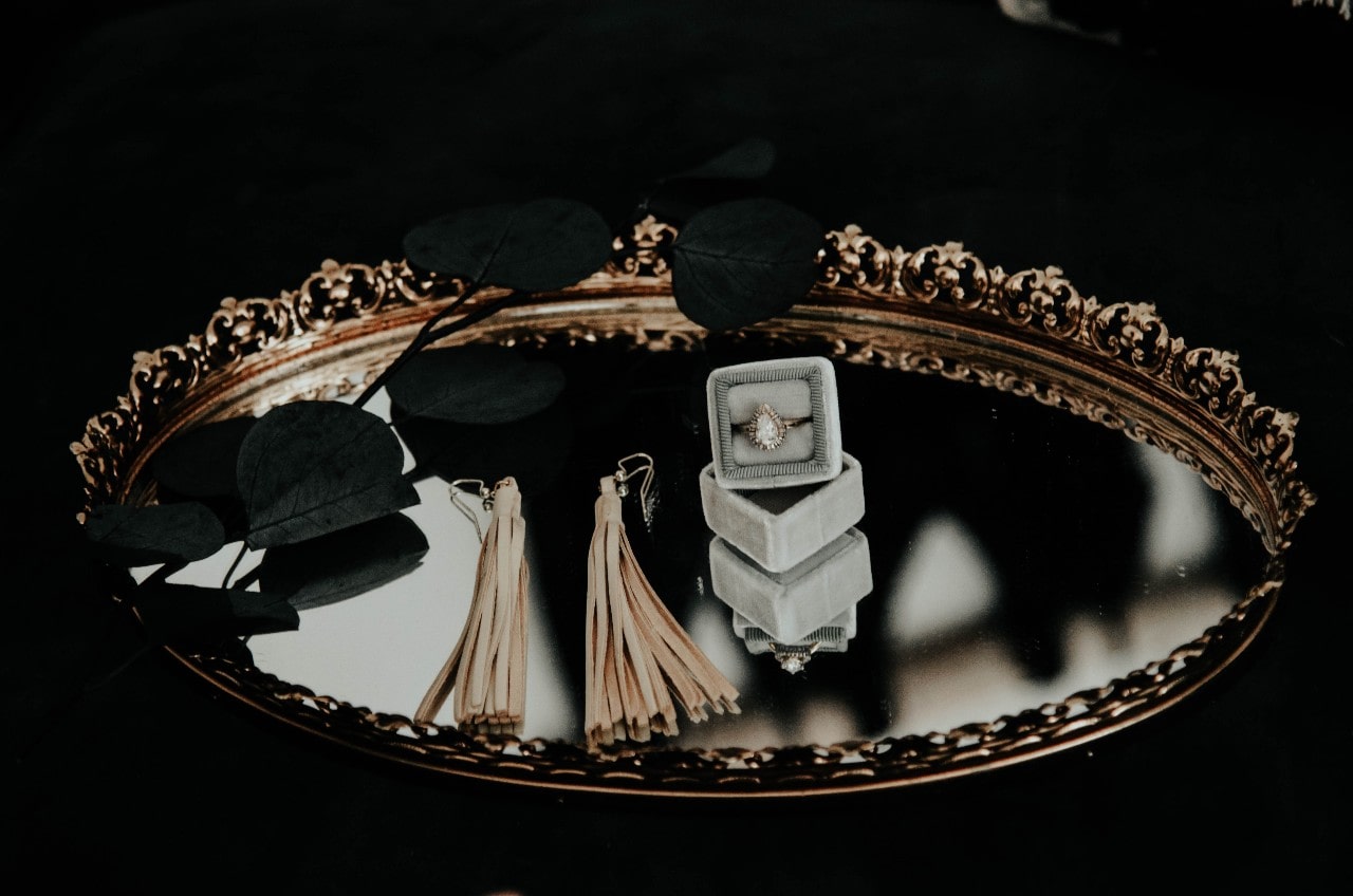 An engagement ring in a box sits on a tray with tassel earrings and black leaves. An engagement ring in a box sits on a tray with tassel earrings and black leaves.