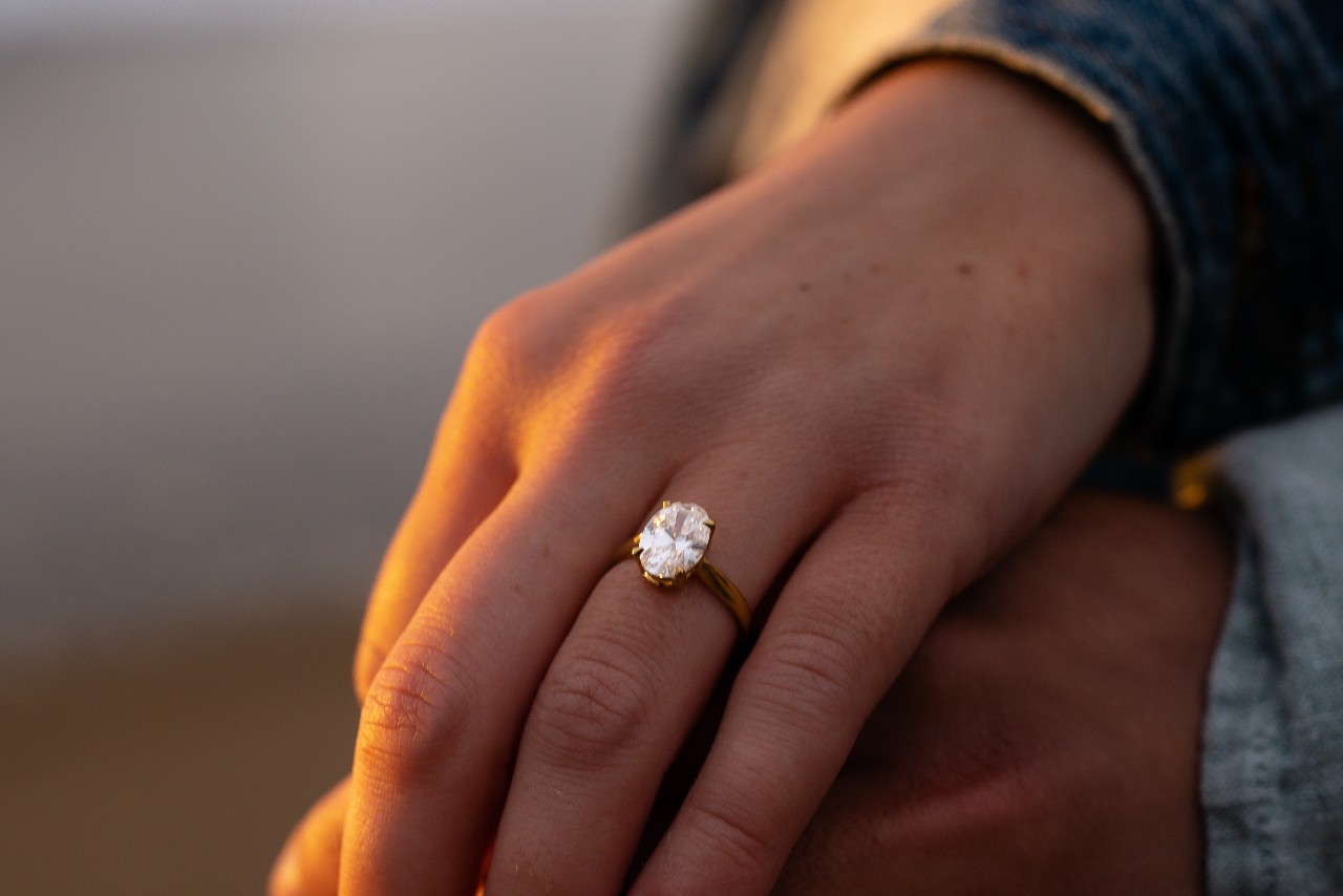 A woman’s hand wearing an oval solitaire ring sits on top of her partner’s hand.