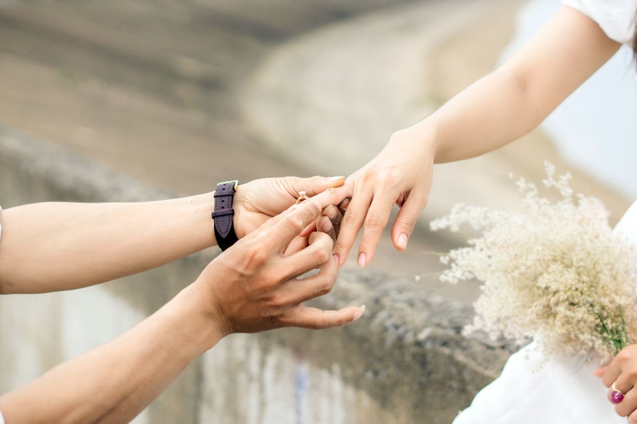 A man slips an engagement ring on the finger of a bride.