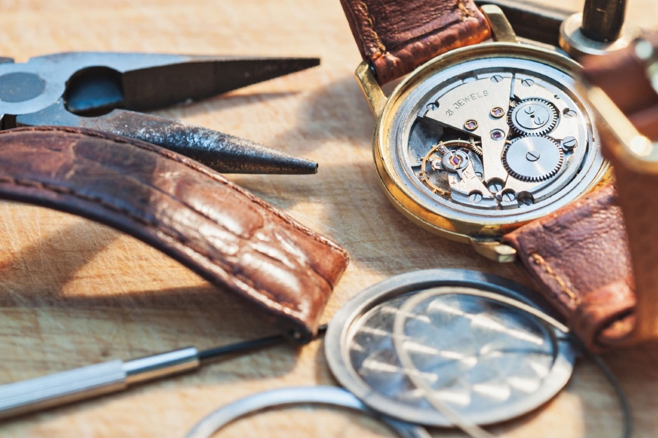 Open watch with visible gears and "21 JEWELS" engraving on a wooden table. Brown leather strap, pliers, and tools nearby, suggesting repair work. Open watch with visible gears and "21 JEWELS" engraving on a wooden table. Brown leather strap, pliers, and tools nearby, suggesting repair work.