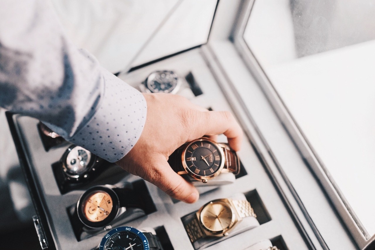 A hand in a polka dot shirt selects a gold watch from a display case filled with various timepieces. A hand in a polka dot shirt selects a gold watch from a display case filled with various timepieces.