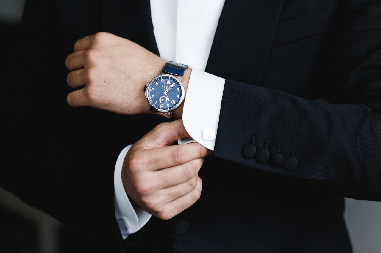A person in a black suit adjusts their silver watch with a blue dial. A person in a black suit adjusts their silver watch with a blue dial.