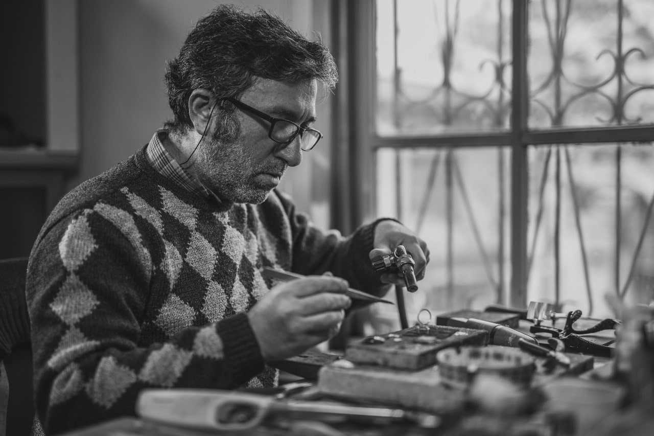A craftsman wearing glasses carefully works on jewelry with tools at a cluttered workbench beside a window.