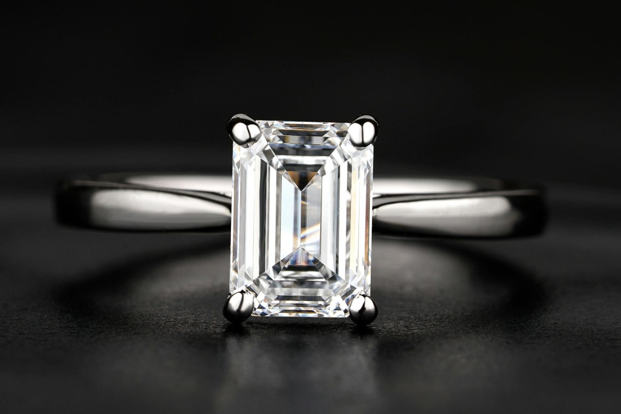 A close-up of a simple yet stunning emerald cut engagement ring on a black backdrop. A close-up of a simple yet stunning emerald cut engagement ring on a black backdrop.