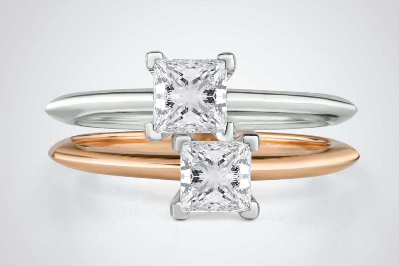A close-up of two cushion cut solitaire rings on a pale gray background. A close-up of two cushion cut solitaire rings on a pale gray background.